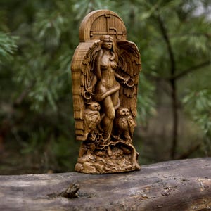 May include: A wooden carving of a winged female figure with a snake wrapped around her. She is standing on a base with two owls on either side. The carving is in a natural wood color.