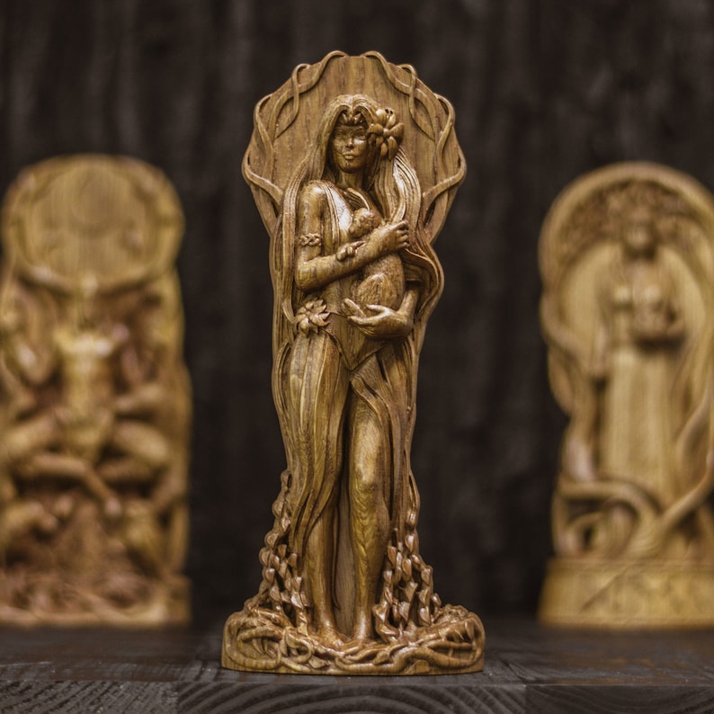 Puede incluir: Una escultura de madera de una mujer sosteniendo un animal peque&ntilde;o en sus brazos. La mujer est&aacute; tallada con un vestido fluido con detalles intrincados. La escultura est&aacute; rodeada de hojas y enredaderas talladas.