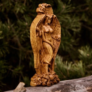 Puede incluir: Una estatua de madera de una figura femenina con alas y una serpiente enroscada a su alrededor. La figura está de pie sobre una base de cráneos. La estatua está tallada en madera marrón oscura y tiene un acabado desgastado.