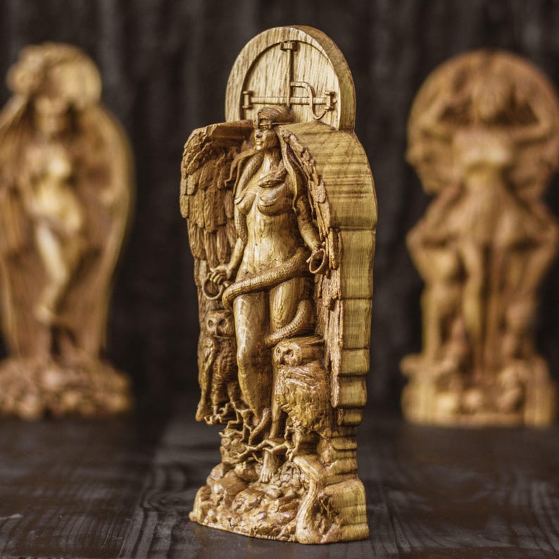 May include: A wooden sculpture of a woman with wings, blindfolded, and holding a snake. The sculpture is carved in a detailed style and features two owls on either side of the woman. The sculpture is set against a dark background.