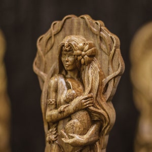 Puede incluir: Una talla de madera de una mujer con el pelo largo y suelto que sostiene un animal peque&ntilde;o en sus brazos. La talla es detallada e intrincada, con un dise&ntilde;o floral alrededor de la cabeza de la mujer.