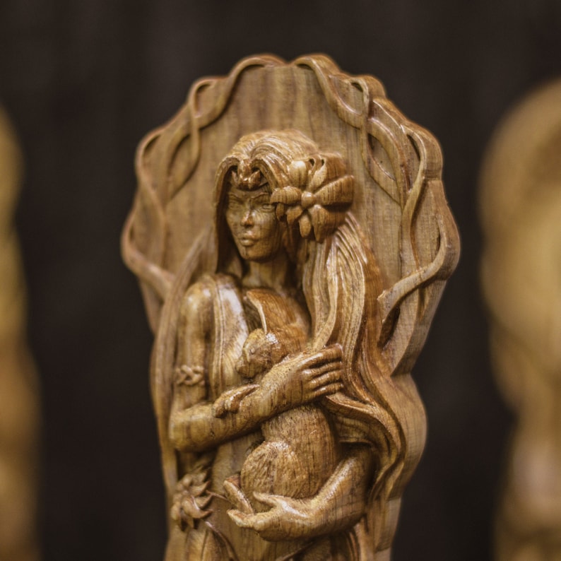 Puede incluir: Una talla de madera de una mujer con el pelo largo y suelto que sostiene un animal peque&ntilde;o en sus brazos. La talla es detallada e intrincada, con un dise&ntilde;o floral alrededor de la cabeza de la mujer.