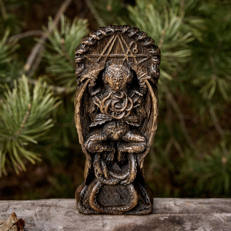 May include: A wooden statue of a creature with wings and a snake wrapped around its torso. The creature is sitting in a throne-like structure with a pentagram design. The statue is dark brown and has a weathered look.