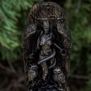 Puede incluir: Una talla de madera oscura de una figura femenina con alas y una serpiente enroscada a su alrededor. La figura est&aacute; de pie sobre una base con dos b&uacute;hos a cada lado. La talla es detallada e intrincada.