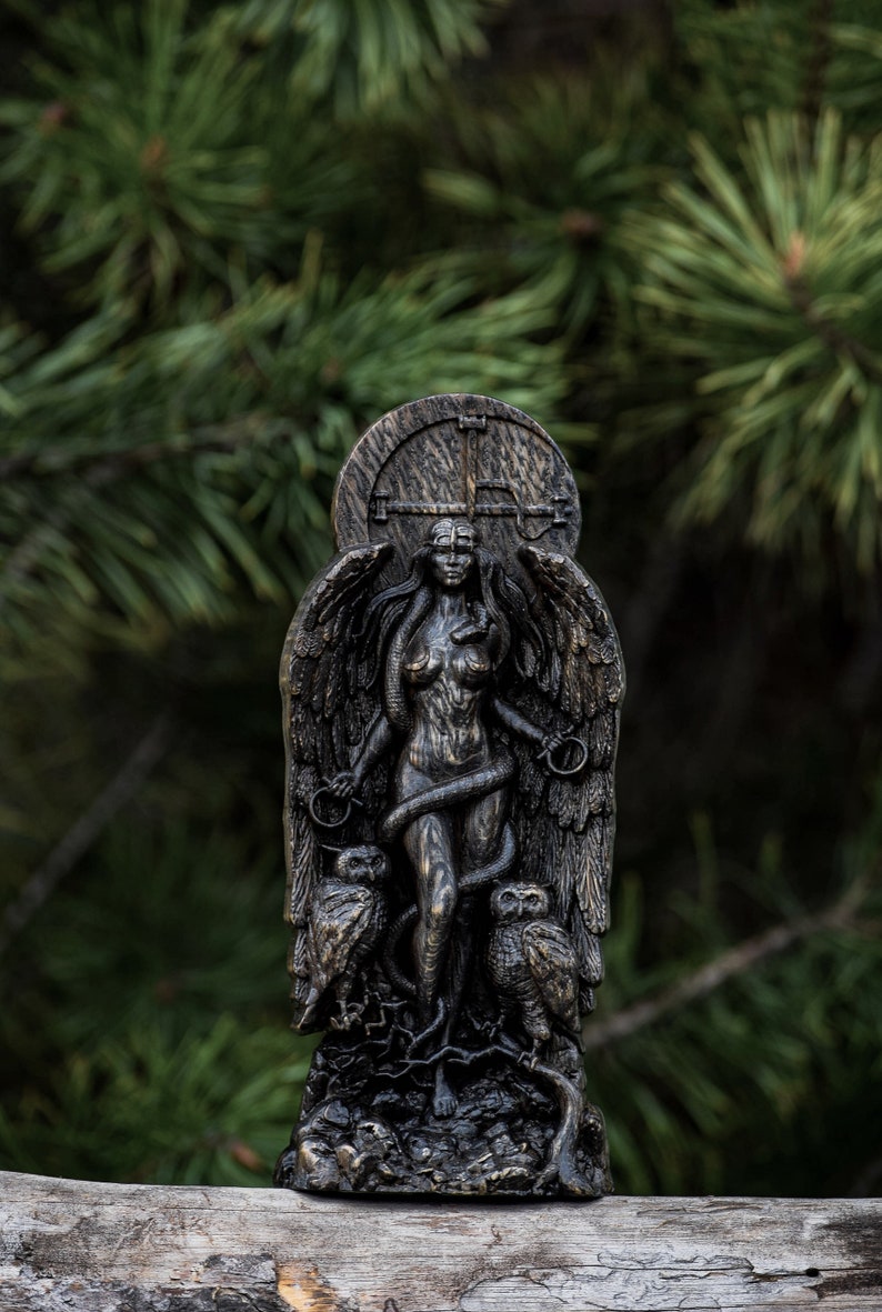 Puede incluir: Una estatua de madera oscura de una mujer alada con una serpiente enroscada a su alrededor. Ella est&aacute; de pie sobre una base con dos b&uacute;hos a cada lado. La estatua est&aacute; colocada sobre un fondo borroso de agujas de pino verdes.