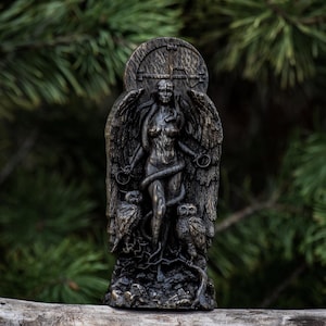Puede incluir: Una estatua de madera oscura de una figura femenina alada con una serpiente enroscada a su alrededor. Ella est&aacute; de pie sobre una base con dos b&uacute;hos a cada lado. La estatua est&aacute; tallada con intrincados detalles y tiene un aspecto r&uacute;stico y antiguo.
