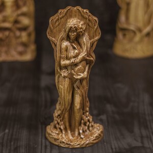Puede incluir: Una escultura de madera de una mujer de pie con una t&uacute;nica fluida, sosteniendo una peque&ntilde;a figura en sus brazos. La escultura est&aacute; tallada con intrincados detalles y tiene un acabado de madera natural.