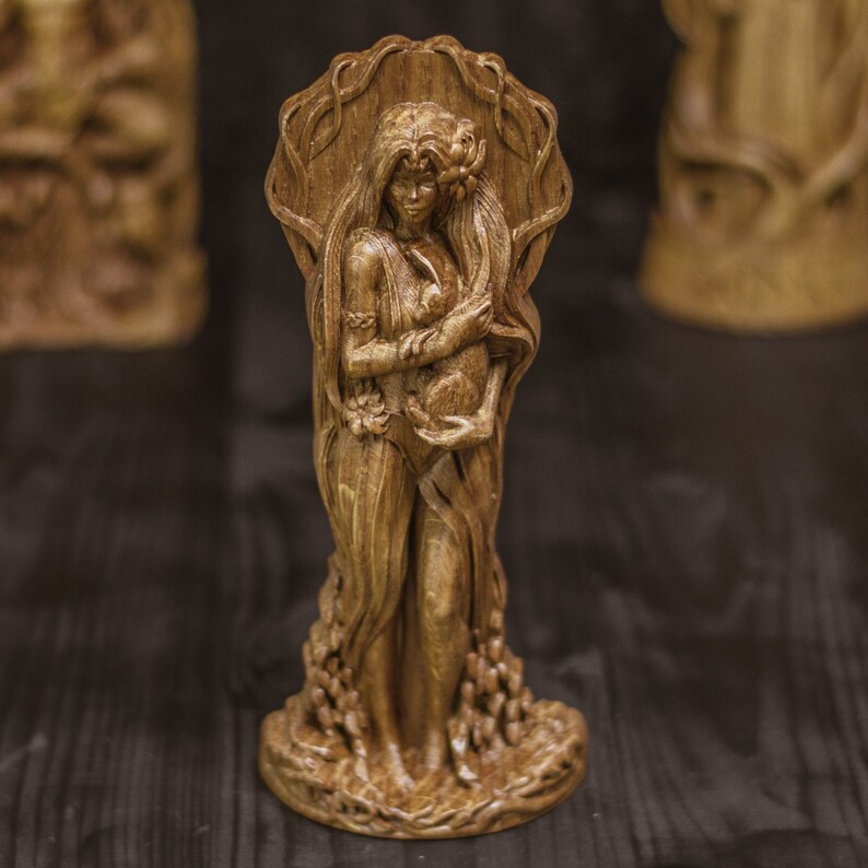 Puede incluir: Una escultura de madera de una mujer de pie con una t&uacute;nica fluida, sosteniendo una peque&ntilde;a figura en sus brazos. La escultura est&aacute; tallada con intrincados detalles y tiene un acabado de madera natural.