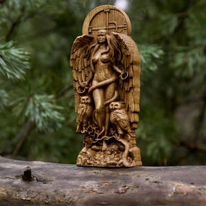 May include: A wooden carving of a winged female figure with a snake wrapped around her. The figure is standing in front of a carved wooden door with two owls on either side. The carving is painted in shades of brown and has a rustic appearance.