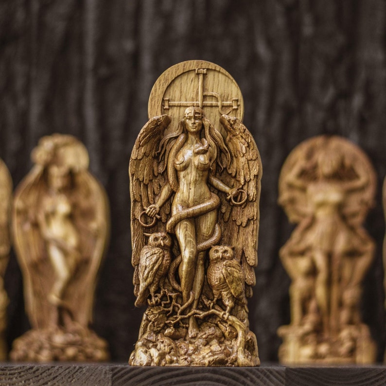 May include: A wooden carving of a winged female figure with a snake wrapped around her. She is standing on a base with two owls on either side. The carving is painted in shades of brown and gold.