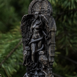 Puede incluir: Una talla de madera oscura de una figura femenina con alas, una serpiente y búhos. La figura está de pie frente a una puerta de madera con un diseño en forma de cruz.