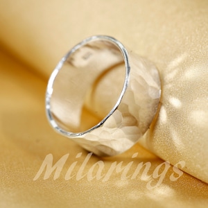 Op de afbeelding: Een zilveren ring met een gehammerde textuur. De ring is breed en heeft een afgeronde vorm.