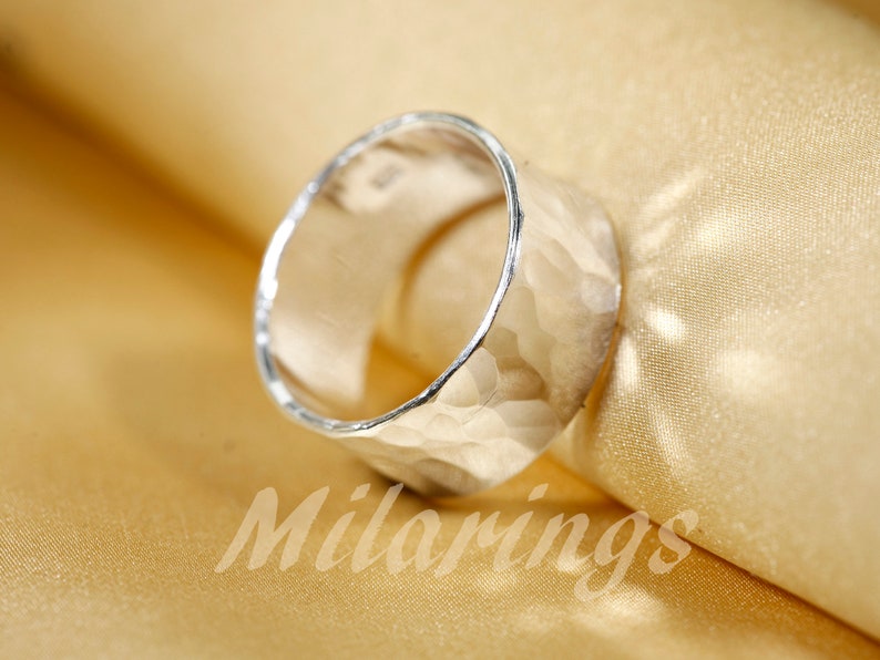 Op de afbeelding: Een zilveren ring met een gehammerde textuur. De ring is breed en heeft een afgeronde vorm.