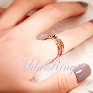 Puede incluir: Un anillo de oro rosa con un dise&ntilde;o de varias bandas, una fila central de peque&ntilde;as piedras redondas oscuras y bordes texturizados. El texto "Mila Rings" est&aacute; impreso en azul en la parte inferior de la imagen.