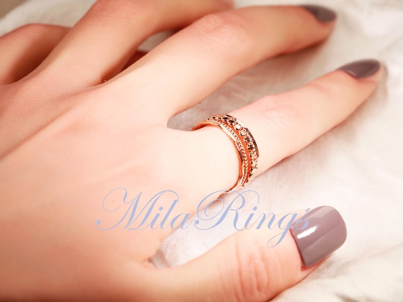 Puede incluir: Un anillo de oro rosa con un dise&ntilde;o de varias bandas, una fila central de peque&ntilde;as piedras redondas oscuras y bordes texturizados. El texto "Mila Rings" est&aacute; impreso en azul en la parte inferior de la imagen.