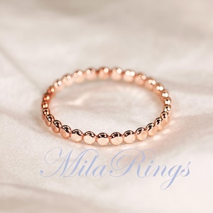 Puede incluir: Un anillo de oro rosa que presenta una banda continua de peque&ntilde;as cuentas esf&eacute;ricas uniformes dispuestas en un patr&oacute;n circular. El texto "Mila Rings" est&aacute; impreso en una fuente cursiva azul claro en la parte inferior de la imagen, centrado.