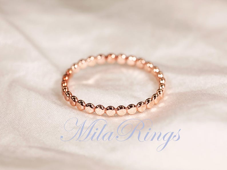 Puede incluir: Un anillo de oro rosa que presenta una banda continua de peque&ntilde;as cuentas esf&eacute;ricas uniformes dispuestas en un patr&oacute;n circular. El texto "Mila Rings" est&aacute; impreso en una fuente cursiva azul claro en la parte inferior de la imagen, centrado.