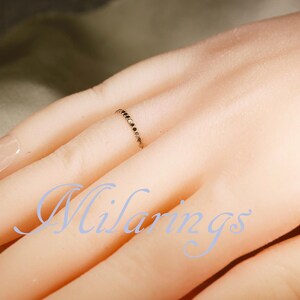 May include: A delicate gold ring with small black stones is worn on a finger. The ring is thin and elegant, with the stones set closely together. The image also shows a hand with a French manicure.