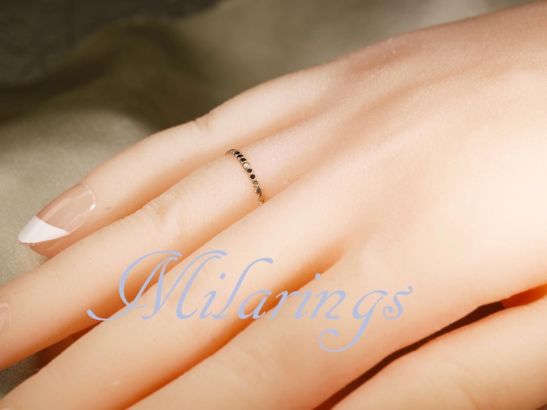 May include: A delicate gold ring with small black stones is worn on a finger. The ring is thin and elegant, with the stones set closely together. The image also shows a hand with a French manicure.