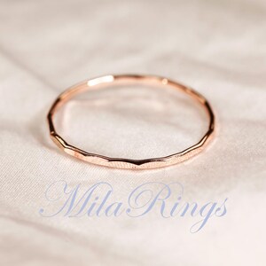 Puede incluir: Un anillo fino de oro rosa con una textura facetada y martillada a lo largo de la banda exterior. El anillo es circular y presenta un acabado met&aacute;lico. El texto "Mila Rings" est&aacute; impreso en una fuente cursiva azul claro en la parte inferior.