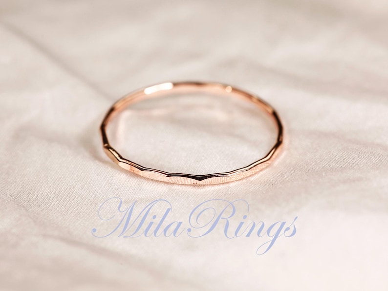Puede incluir: Un anillo fino de oro rosa con una textura facetada y martillada a lo largo de la banda exterior. El anillo es circular y presenta un acabado met&aacute;lico. El texto "Mila Rings" est&aacute; impreso en una fuente cursiva azul claro en la parte inferior.
