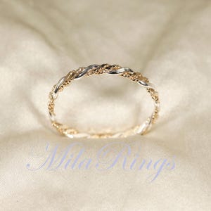 May include: A two-tone ring with a twisted rope design. The ring features alternating sections of gold and silver metal, creating a textured and elegant appearance. The ring is displayed on a soft, cream-colored fabric.