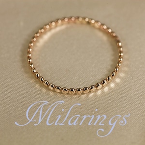 May include: A delicate gold ring with a beaded design. The ring is circular and sits on a cream-colored surface. The word "Milarings" is written in cursive script below the ring.