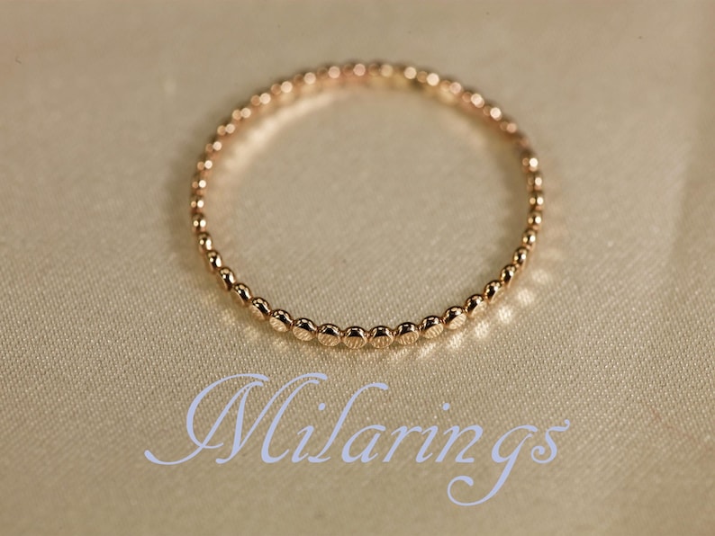 May include: A delicate gold ring with a beaded design. The ring is circular and sits on a cream-colored surface. The word "Milarings" is written in cursive script below the ring.