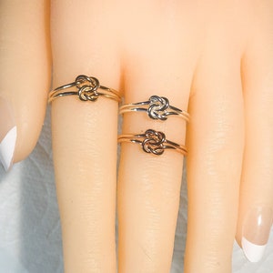 Op de afbeelding: Drie gouden knoop ringen op een hand. De ringen zijn op elkaar gestapeld, met de grootste ring onderaan en de kleinste ring bovenaan.