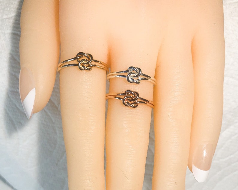 Op de afbeelding: Drie gouden knoop ringen op een hand. De ringen zijn op elkaar gestapeld, met de grootste ring onderaan en de kleinste ring bovenaan.