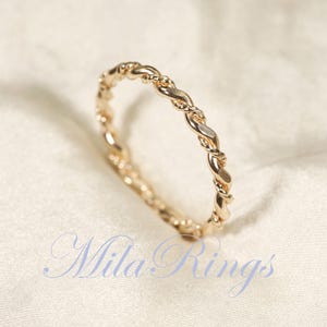 Peut inclure: Une bague dor&eacute;e avec un motif de corde torsad&eacute;e. La bague est pos&eacute;e sur un fond blanc cass&eacute;. Le texte "Mila Rings" est visible dans la partie inf&eacute;rieure de l'image.