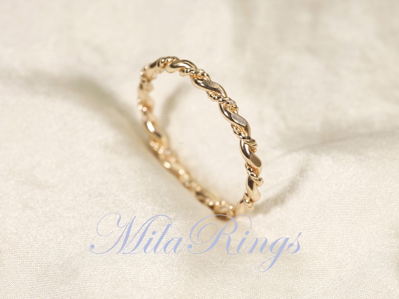 Peut inclure: Une bague dor&eacute;e avec un motif de corde torsad&eacute;e. La bague est pos&eacute;e sur un fond blanc cass&eacute;. Le texte "Mila Rings" est visible dans la partie inf&eacute;rieure de l'image.