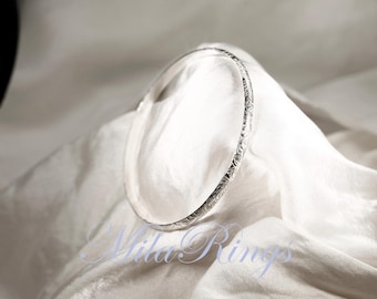 May include: A silver bangle bracelet with a delicate floral design. The bracelet is smooth and shiny, and it is lying on a white fabric background.
