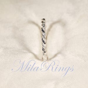 Peut inclure: Une bague argent&eacute;e avec un motif torsad&eacute; en forme de corde. La bague est centr&eacute;e sur un fond de tissu blanc. Le texte "Mila Rings" est affich&eacute; en cursive en bas de l'image.