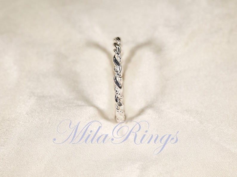 Peut inclure: Une bague argent&eacute;e avec un motif torsad&eacute; en forme de corde. La bague est centr&eacute;e sur un fond de tissu blanc. Le texte "Mila Rings" est affich&eacute; en cursive en bas de l'image.