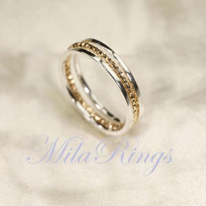 May include: A silver ring with a gold-colored, textured band. The ring has a double-band design, with a smooth silver outer band and a textured inner band. The ring is displayed on a soft, white surface. The text "Mila Rings" is visible.