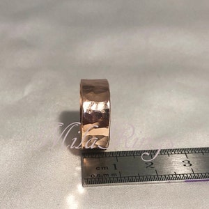 May include: A hammered rose gold ring measuring 1 cm in width. The ring is shown next to a ruler for size reference.