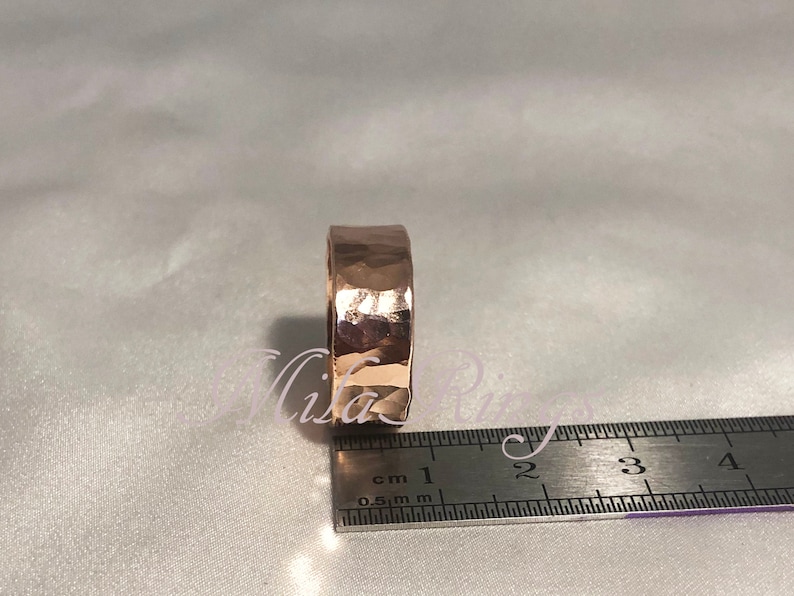May include: A hammered rose gold ring measuring 1 cm in width. The ring is shown next to a ruler for size reference.