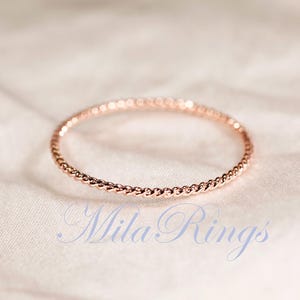 Puede incluir: Un anillo fino de oro rosa que presenta una delicada textura de cuerda trenzada en toda la banda. El texto MilaRings est&aacute; impreso en una fuente cursiva de color azul claro debajo del anillo, situado sobre una superficie de tela suave.