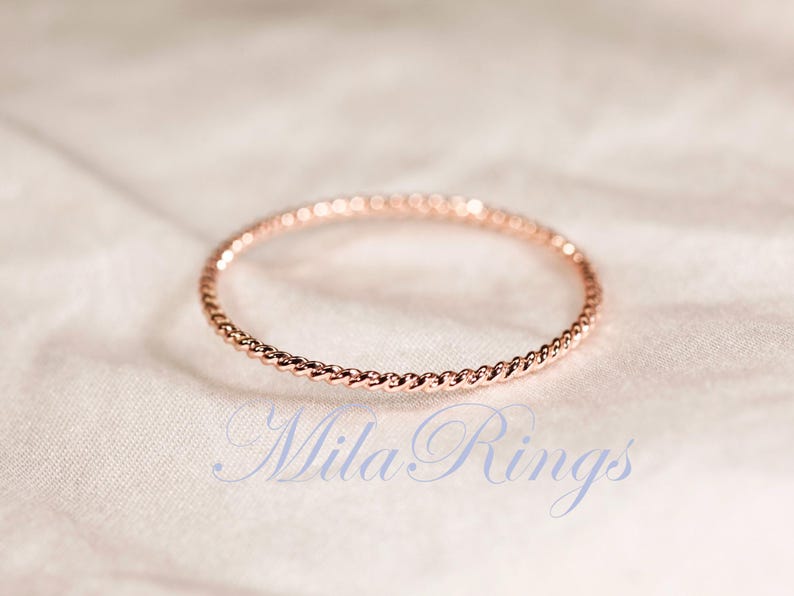 Puede incluir: Un anillo fino de oro rosa que presenta una delicada textura de cuerda trenzada en toda la banda. El texto MilaRings est&aacute; impreso en una fuente cursiva de color azul claro debajo del anillo, situado sobre una superficie de tela suave.