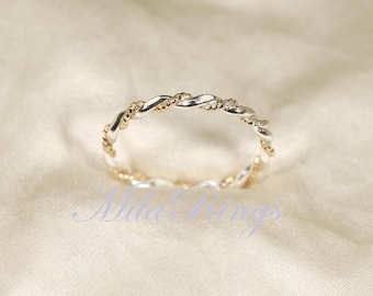May include: A silver and gold-toned twisted rope ring. The ring features a braided design with alternating silver and gold sections. The ring is set against a soft, off-white background. The text "Nila Rings" is visible in the image.