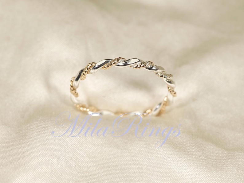 Peut inclure: Une bague torsad&eacute;e argent&eacute;e et dor&eacute;e. La bague pr&eacute;sente un motif tress&eacute; avec des sections argent&eacute;es et dor&eacute;es altern&eacute;es. La bague est pos&eacute;e sur un fond blanc cass&eacute;. Le texte "Nila Rings" est visible dans l'image.