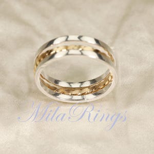 May include: A silver ring with two gold bands. The ring has a textured design on the gold bands. The ring is on a white fabric. The text "Mila Rings" is at the bottom of the image.
