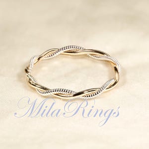 Può includere: Un anello sottile dal design intrecciato con fili alternati in metallo color oro lucido e metallo color argento testurizzato. Il testo Mila Rings è stampato in un carattere corsivo azzurro sotto l'anello sulla superficie chiara.