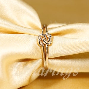May include: A gold ring with a knot design. The ring is on a light brown fabric background.