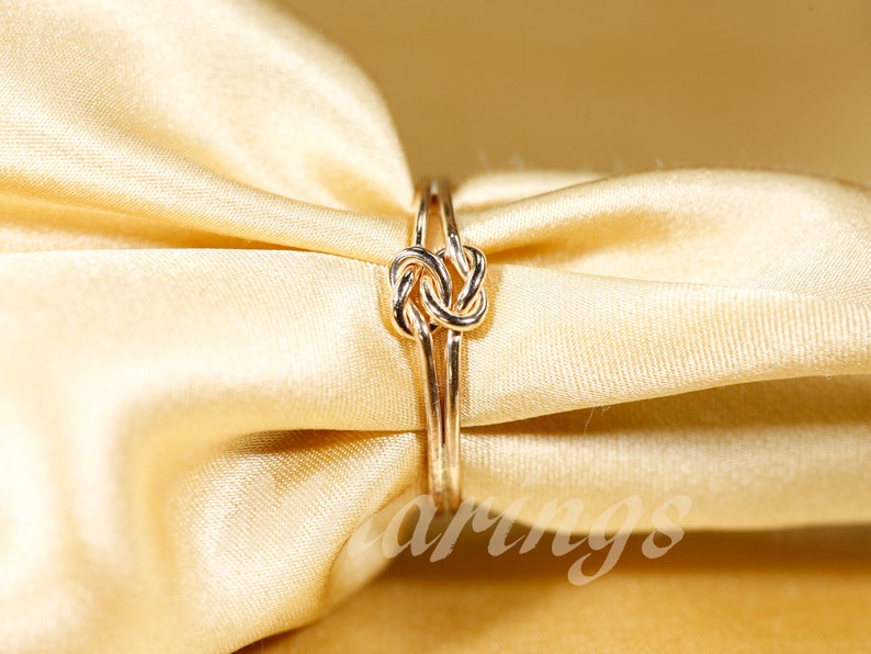 May include: A gold ring with a knot design. The ring is on a light brown fabric background.