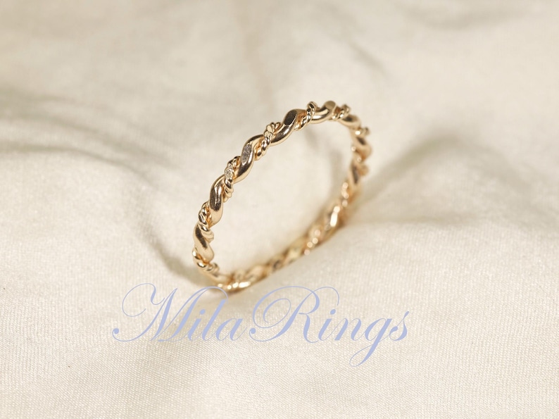 Peut inclure: Une bague dor&eacute;e avec un motif de corde torsad&eacute;e. La bague est en m&eacute;tal brillant et pr&eacute;sente un motif d&eacute;licat et entrelac&eacute;. La bague est un bijou, probablement pour le doigt, et est pr&eacute;sent&eacute;e sur un fond de tissu blanc. Le texte "Mila Rings" est visible.