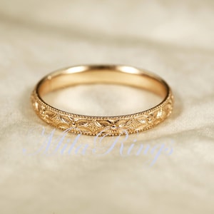 May include: A rose gold band ring featuring a continuous engraved floral pattern around the exterior surface. The metal has a polished finish with delicate milgrain detailing along the top and bottom edges of the band, creating a textured look.