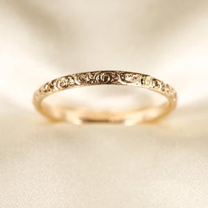 Puede incluir: Un anillo de banda fina de oro con un intrincado patrón de volutas grabado de forma continua en toda su superficie exterior. El metal tiene un acabado pulido y reflectante que capta la luz, resaltando el relieve detallado de los grabados.