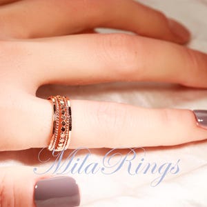 Puede incluir: Un anillo en tono oro rosa con una banda central de peque&ntilde;as piedras circulares oscuras, flanqueada por dos filas de detalles met&aacute;licos texturizados y con cuentas. El anillo tiene un perfil ancho y plano. El texto Mila Rings es visible.